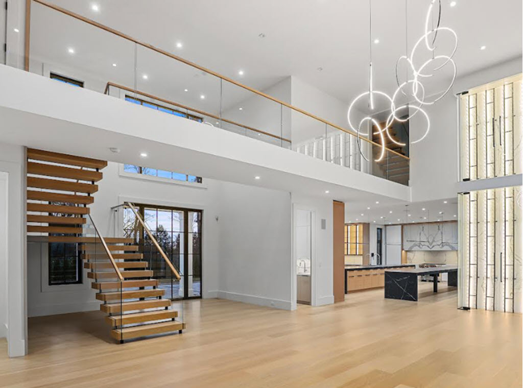 Greenwich home with a large open room featuring a floating double stringer staircse with glass railings, The modern kitchen can be seen in the back ground. A lighted glass wall addes to the modern aesthetic and the modern circle chain light flows down from the ceiling.