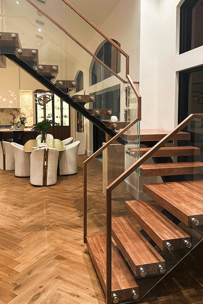 floating mono-stringer staircase with glass railing, and walnut wood treads in  great room with herringbone wood floors and modern decor.